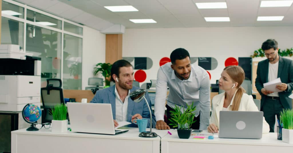 A colleague leans over to assist two others in an open office setting illustrating the idea of sales enablement best practices.