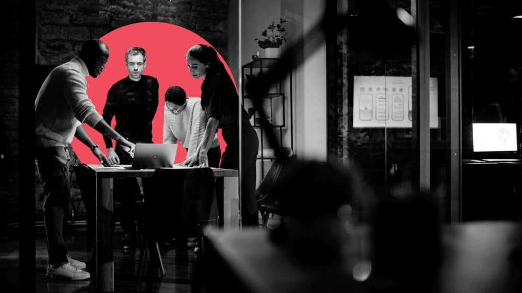 In a dark office, a diverse group of sales and marketing team members discuss alignment on key goals over a laptop.