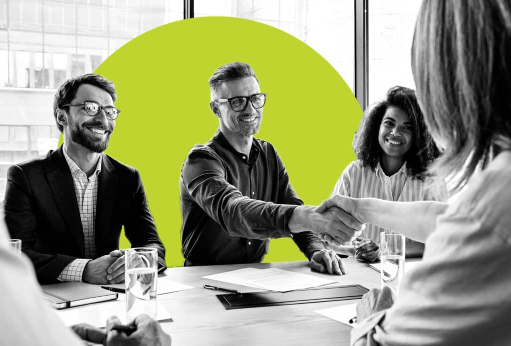 A diverse group of sales people shake hands across a conference table with a marketer in the foreground to illustrate sales and marketing allignment