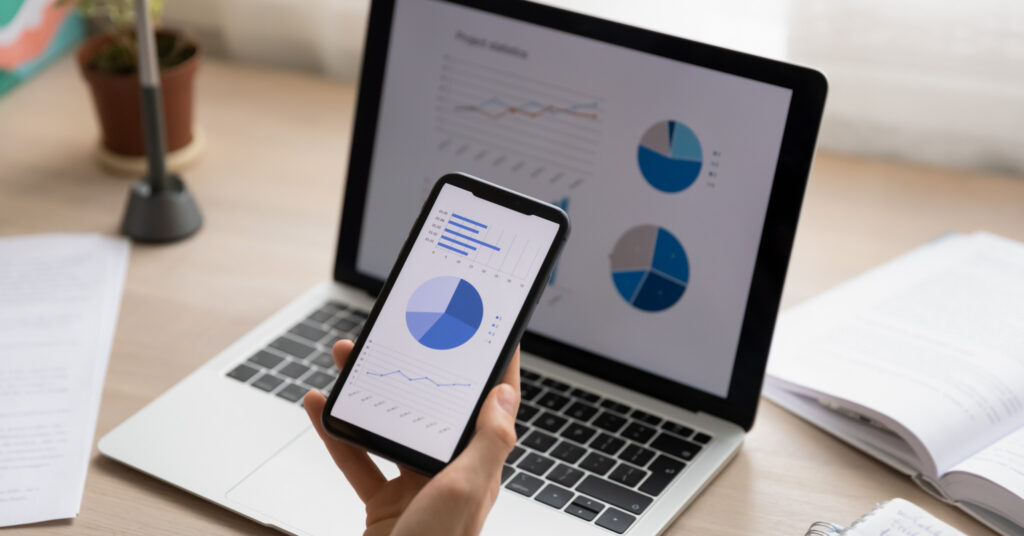A closeup of a white hand holding a smartphone in front of a laptop, both screens show data graphics