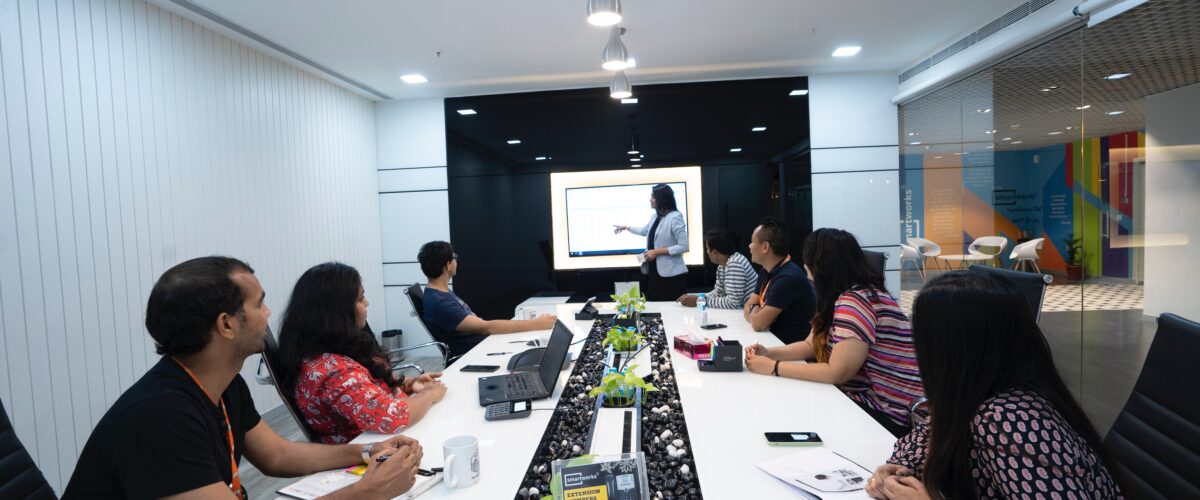 A group of office workers seated at a large conference table watch a presentation on screen
